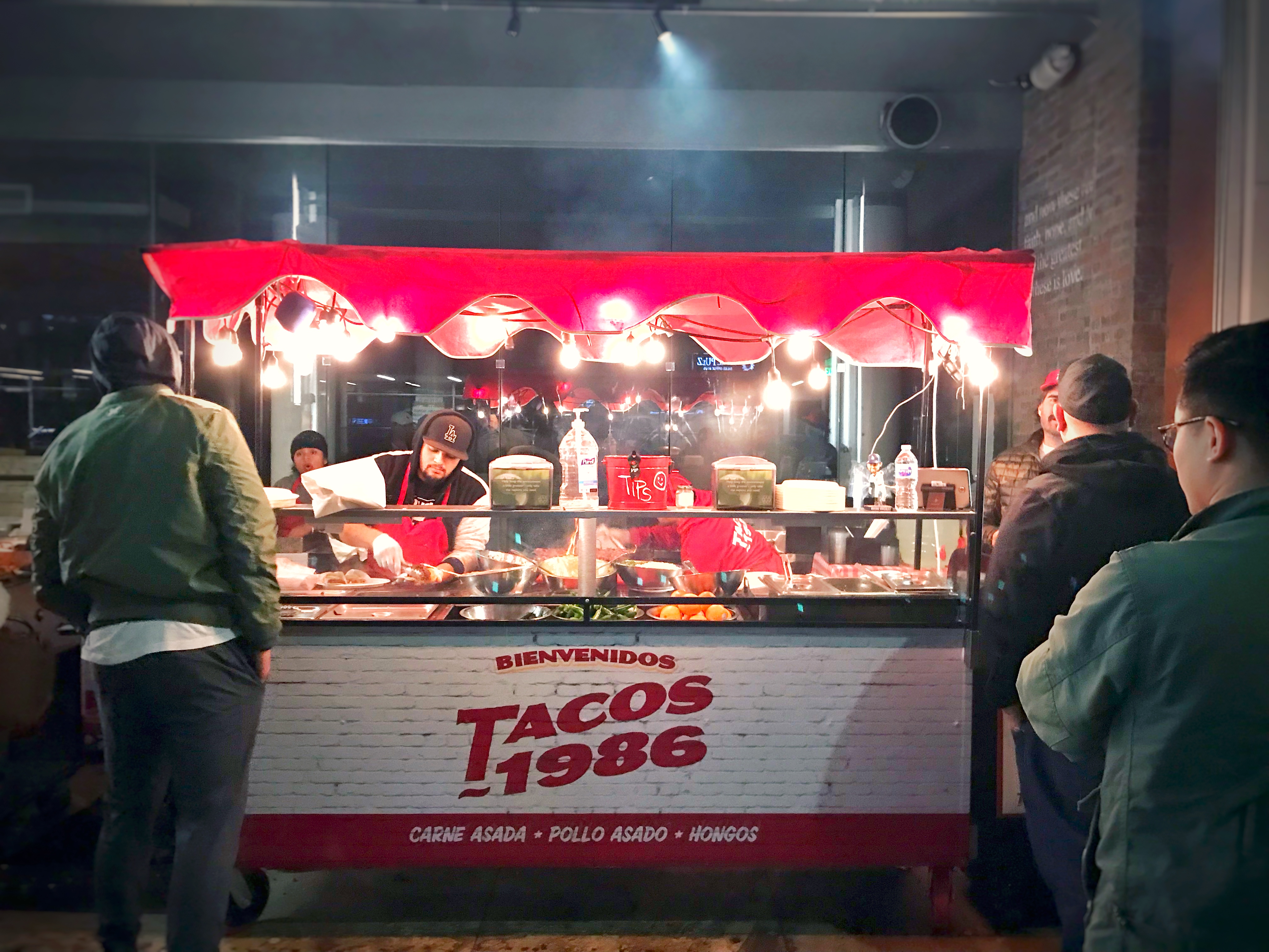 Cooks serving customers at the early Tacos 1986 Koreatown taco stand beneath red awnings